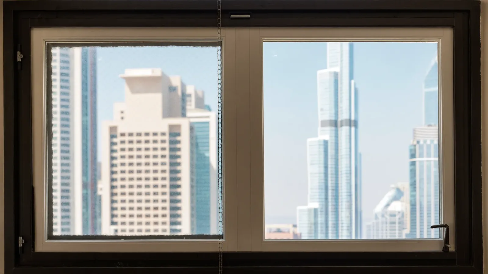 A secondary glazing unit in a slim white frame fitted in front of an existing dark-framed window, showing the air gap between the two panes, with Dubai towers beyond.