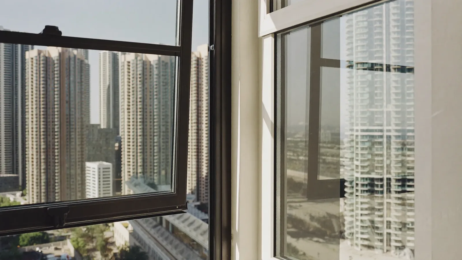A bright Dubai apartment corner where a black aluminium-framed window meets a white uPVC-framed one, both looking out over Marina high-rise towers.