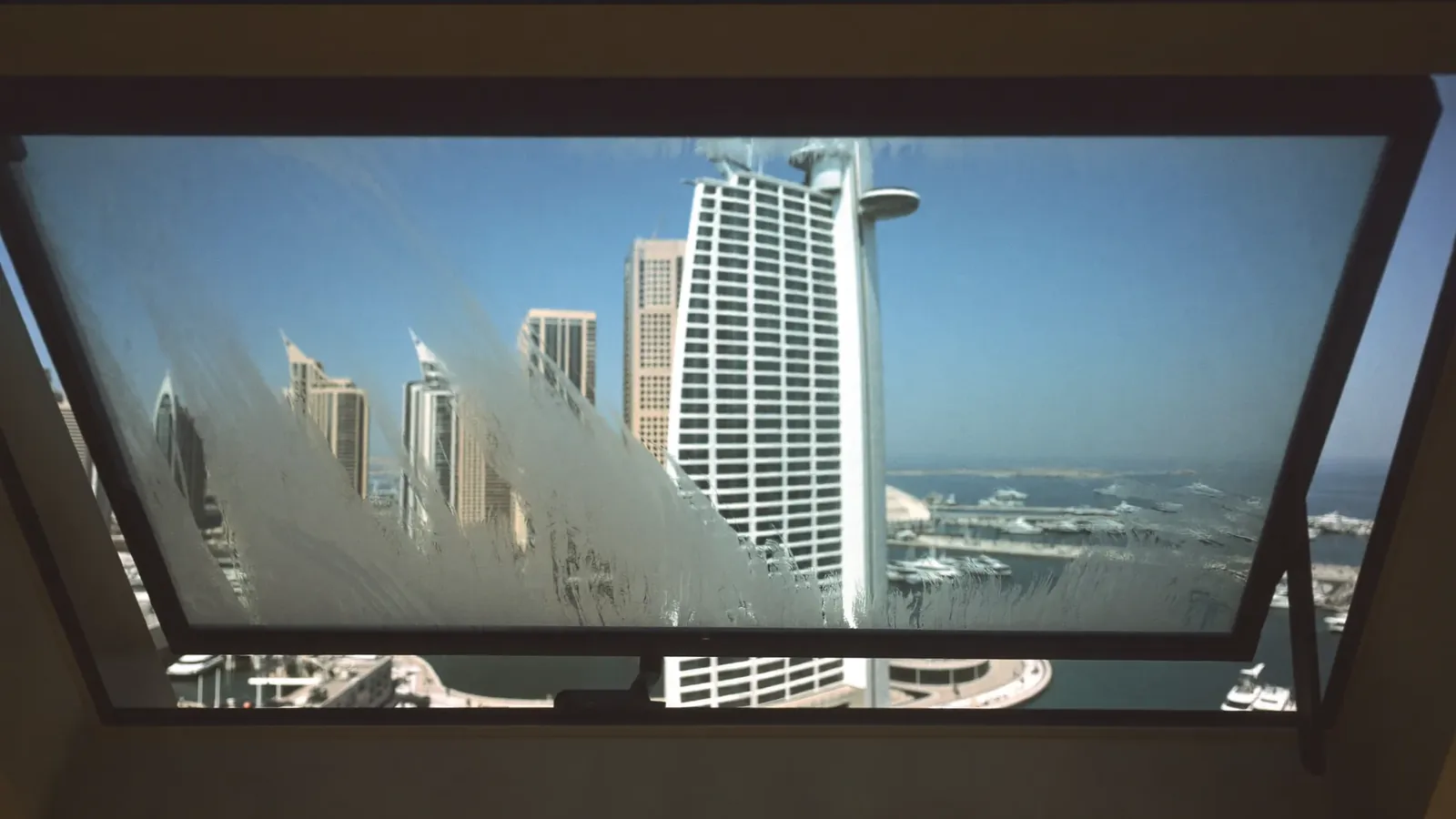 Heavy condensation on the inside of a modern slim-framed apartment window, with a wiped clear streak revealing the Dubai Marina towers, yachts and blue sky beyond.