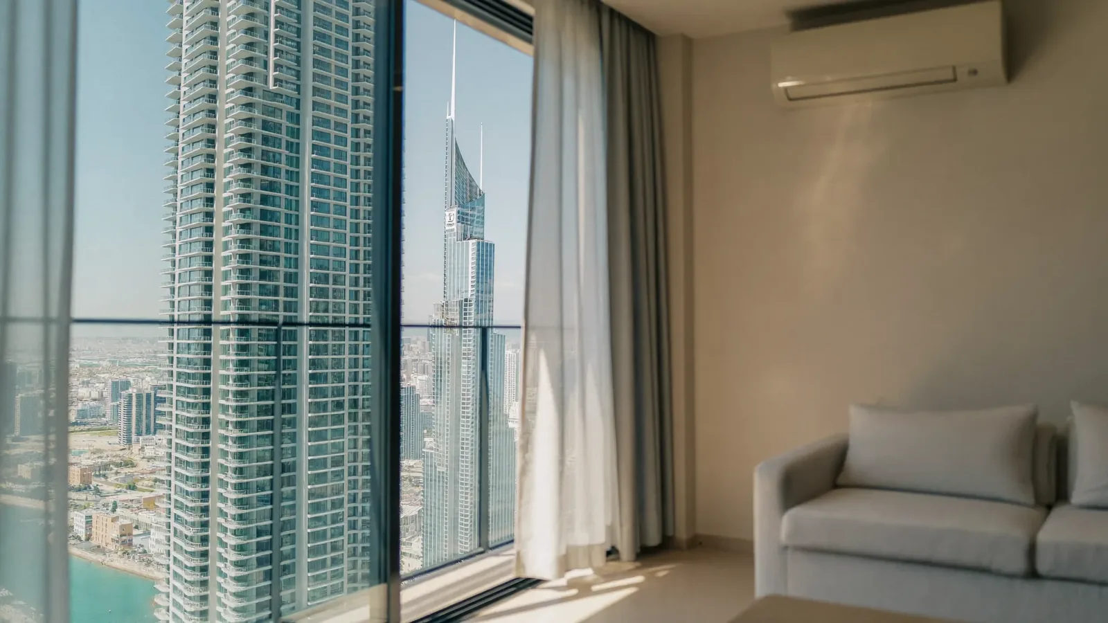 A sunlit Dubai apartment living room with a floor-to-ceiling sliding glass door, one tinted solar-control panel closed against the Marina towers and a wall-mounted air conditioner above a pale linen sofa.