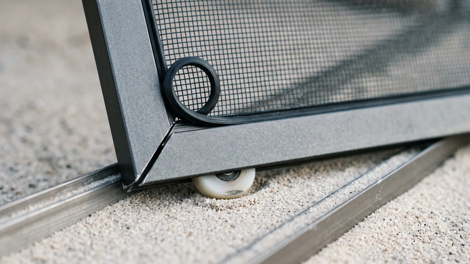Macro of the bottom corner of a mosquito screen panel: an opened mitre joint in the thin aluminium frame, a loop of rubber spline lifted out of its groove, a small nylon roller with a flat worn spot, and the shallow channel below packed level with fine pale sand.