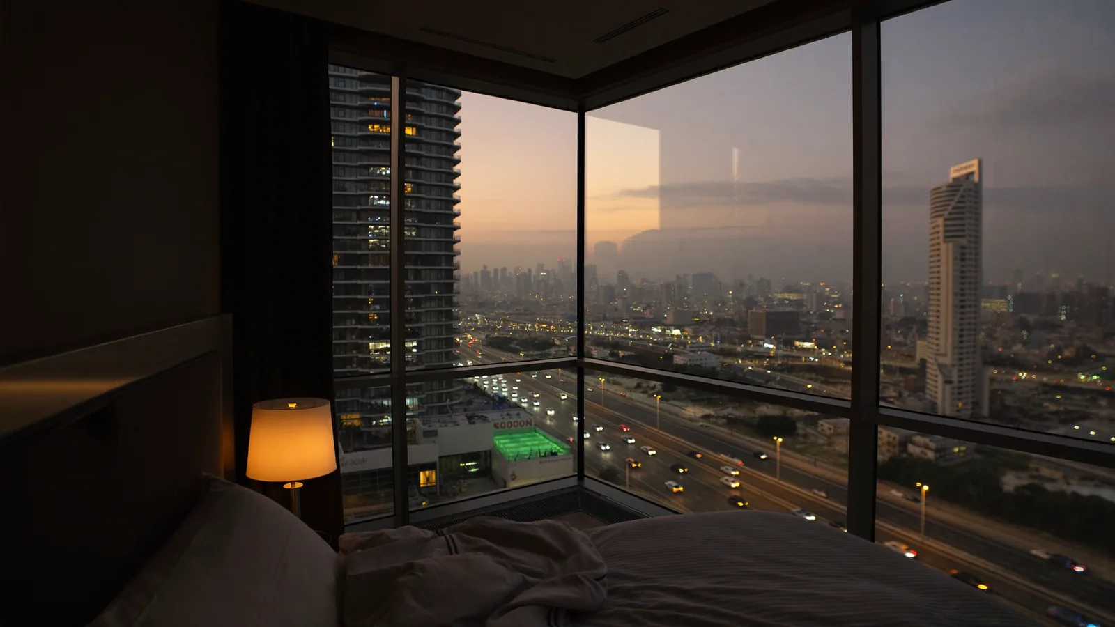 Bedroom at dusk with a panoramic window onto a lit Dubai motorway and the skyline