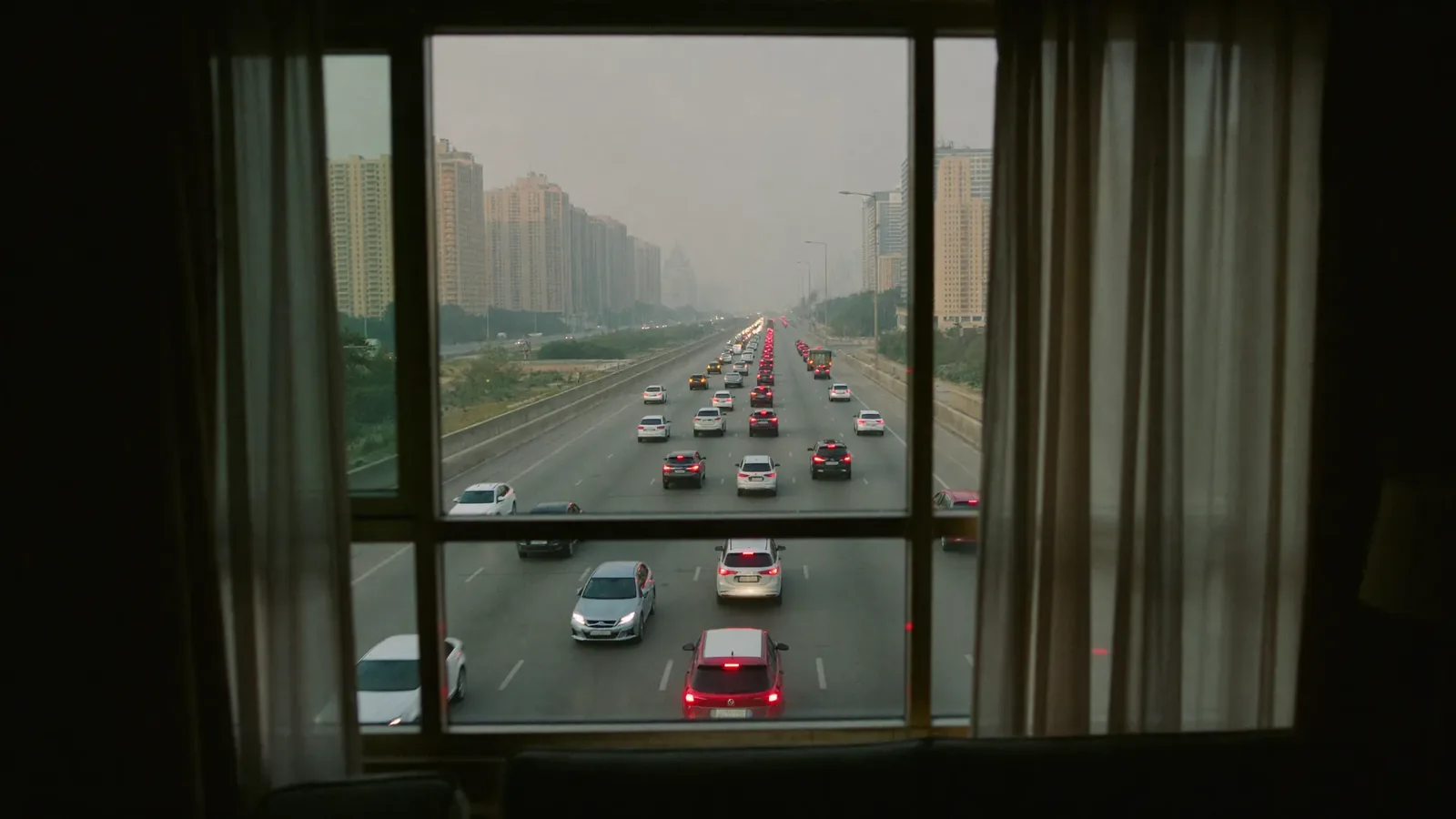A quiet dimly lit apartment living room at dusk behind closed floor-to-ceiling windows, with traffic streaming along a lit multi-lane Dubai highway outside.